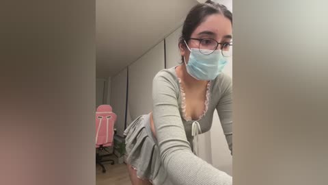 Media: Video of a young woman with light skin and glasses, wearing a light gray dress and a blue surgical mask, leaning forward in a dimly lit room with a pink gaming chair in the background.