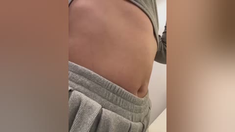 Media: Close-up video of a person's bare midriff and lower torso, showing light skin with a slight tan, wearing grey sweatpants pulled down to reveal a toned abdomen. Background is out of focus.