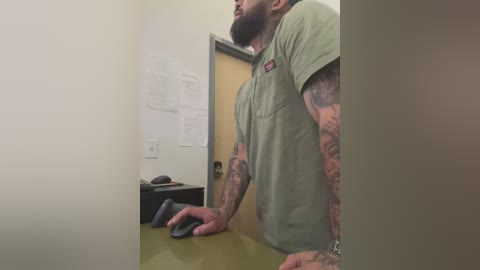 Media: Video of a tattooed man with a beard, wearing a green t-shirt, leaning on a desk, in a dimly lit office with beige walls and a door.