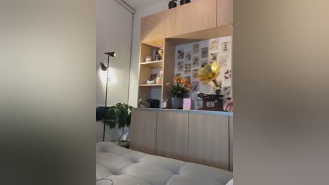 Media: Video of a modern living room with beige walls, light wood cabinets, a tufted sofa, and a vibrant yellow balloon. The room is well-lit by a tall floor lamp and has a cozy, minimalist decor.