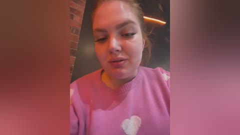 Media: Video of a young woman with fair skin, brown hair, and light pink sweater, indoors with brick wall and blurred background.