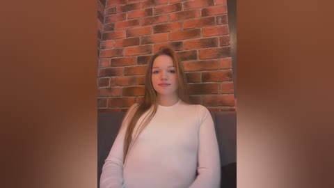 Media: Video of a young pregnant Caucasian woman with long straight hair, wearing a white long-sleeved shirt, standing against a brick wall background.