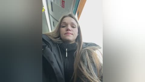 Media: A video of a young woman with long, straight blonde hair, wearing a black, high-necked sweater, sitting in a subway car. Her expression is neutral. Background shows subway doors and a red sign.