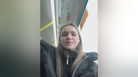 Media: Video of a young woman with long, straight brown hair, wearing a black coat, standing in a subway car. The background shows yellow handles and a bright, white, reflective ceiling.