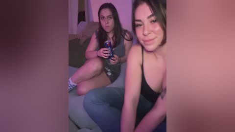 Media: A video of two young women in a dimly lit room, one sitting on a couch, the other kneeling, both wearing casual clothing, with a purple hue casting over the scene.