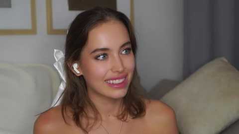 Media: Video of a smiling young woman with light skin and long brown hair, wearing white earbuds, sitting on a beige sofa.