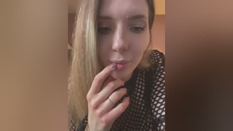 Media: A video shows a young Caucasian woman with straight, light brown hair, wearing a black fishnet top, seductively biting her finger, in a dimly lit room with warm brown walls.