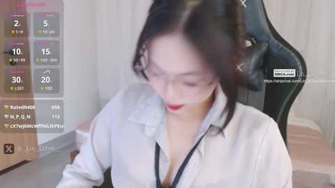 Media: A video of a young Asian woman with fair skin and long black hair, wearing a white blouse with a black collar, sitting at a gaming chair in a dimly lit room.