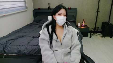 Media: Video of an East Asian woman with long black hair, wearing a white mask, gray hoodie, and sitting on a black chair in a minimalistic bedroom with a dark bed and neutral-colored walls.