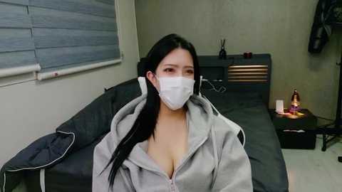 Media: Video of a young Asian woman with long black hair, wearing a white face mask, grey hoodie, and sitting on a dark grey couch in a sparsely decorated room.