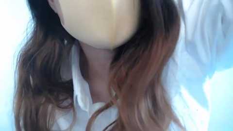 Media: A close-up video of a woman with long, wavy brown hair wearing a white shirt, her face obscured by a beige mask.