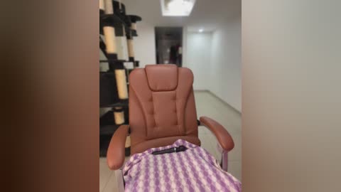 Media: Video of a brown leather massage chair with a purple and white striped blanket draped over the backrest, situated in a modern, minimalist spa room with beige walls, a black shelving unit, and a white ceiling light.