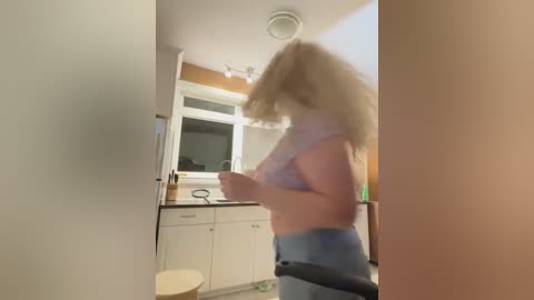 Media: Video of a blonde woman in a white tank top and blue jeans, holding a black object, standing in a dimly lit, beige-walled bathroom with white cabinets and a mirror.