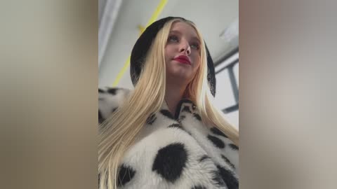 Media: Video of a blonde woman with fair skin, wearing a black hat and a fur coat with black spots, standing indoors with a blurred background.