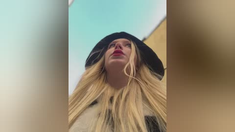 Media: Video of a blonde woman with fair skin, wearing a black beanie and red lipstick, looking up at the sky, with a blurred background.