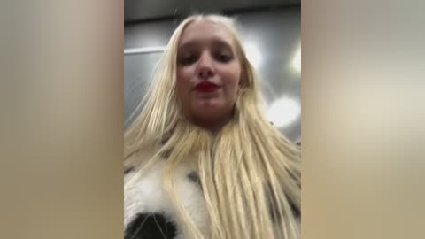 Media: Video of a young, fair-skinned, blonde woman with a full head of hair, styled in a white and black cow pattern. She wears red lipstick. Background is blurred, showing a modern, possibly indoor setting.