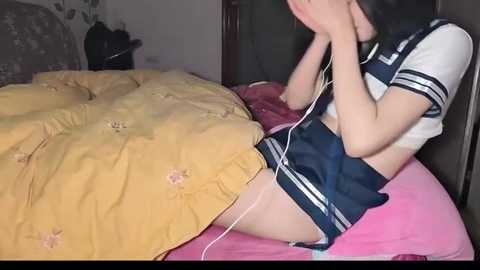 Media: Video of a young Asian woman with fair skin, in a sailor schoolgirl outfit, lying on a bed, listening to music with white earbuds, covered by a yellow blanket.