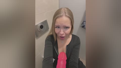 Media: Video of a blonde woman in a public restroom, looking surprised while holding a pink dildo. She wears a black top.