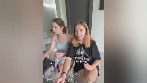 Media: Video of two young women with light skin, one in a blue sports bra, the other in a black t-shirt, sitting on a glass table in a modern, minimalist room.