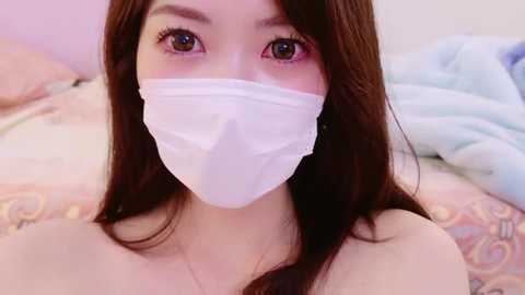 Media: Video of an Asian woman with long brown hair and light skin, wearing a white surgical mask, lying on a bed with pastel-colored bedding, eyes looking directly at the camera.