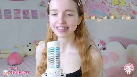 Media: Video of a young, fair-skinned woman with long, wavy blonde hair, wearing a black spaghetti strap top, smiling into a white microphone. Background features a pink and white bedroom with plush toys and a fairy light string.