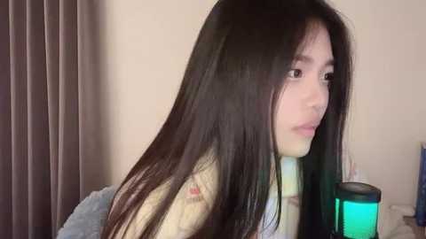 Media: Video of a young Asian woman with long, straight black hair, wearing a white sweater, looking contemplatively to the right. Background includes beige walls and a blue blanket, with a teal light source.