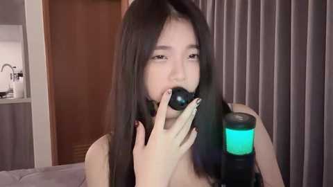 Media: A video of an Asian woman with long black hair, wearing a black strapless top, sucking on a black and turquoise sex toy, in a modern bedroom with beige curtains and a wooden door.