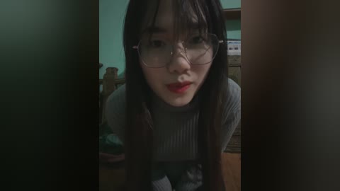 Media: Video of an Asian woman with long black hair and glasses, wearing a grey sweater, leaning forward in a dimly lit room with wooden furniture and green walls.