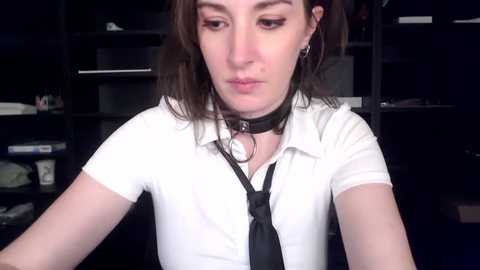 Media: Video of a young woman with fair skin, shoulder-length brown hair, wearing a white collared shirt, black tie, and a black choker, sitting in a dimly lit room with dark bookshelves in the background.