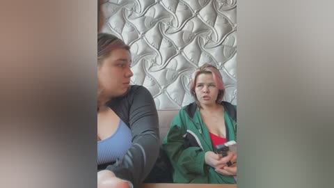 Media: Video of two women sitting in a diner with a tiled wall background. One wears a blue top, the other a green jacket, both holding cups.