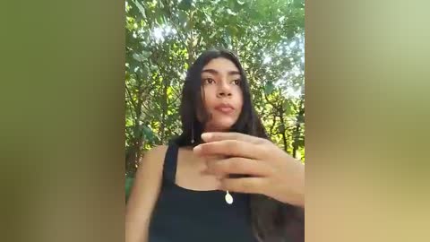 Media: Video of a young woman with long, dark hair, wearing a black tank top, holding a smartphone, standing outdoors amidst lush green foliage.