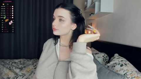 Media: Video of a fair-skinned woman with long black hair, wearing a beige jacket, holding a lit candle, in a cozy, dimly lit bedroom with floral-patterned bedding and a black curtain in the background.