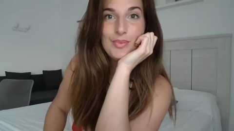 Media: Video of a fair-skinned woman with long, straight brown hair, resting her chin on her hand. She has green eyes and is smiling softly. Background shows a minimalist bedroom with white walls, a bed, and a dark couch.