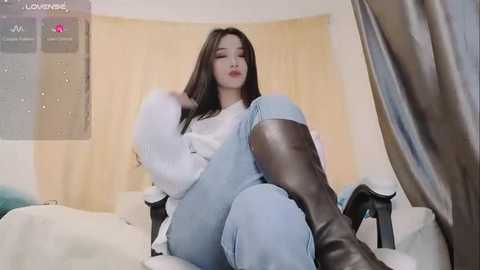 Media: A video of an East Asian woman with long black hair, wearing a white sweater, light blue jeans, and black boots, sitting on a white couch with beige curtains in the background.