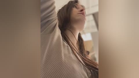 Media: Video of a young woman with long brown hair, wearing a striped sweater, looking upward with a contemplative expression. The background is blurred, suggesting an indoor setting.