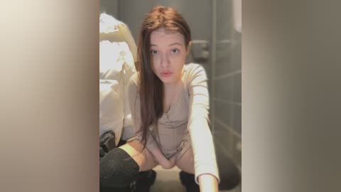 Media: Video of a young Asian woman with fair skin and long brown hair, wearing a beige sweater and black thigh-high socks, squatting in a dimly lit, gray-tiled bathroom, looking directly at the camera.