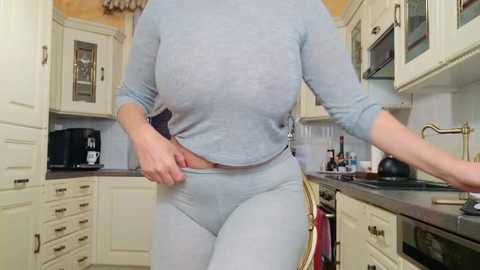 Media: Video of a fair-skinned woman in light blue, long-sleeved, fitted pajamas adjusting her waistband in a cream-colored kitchen with beige cabinets, stainless steel appliances, and wine bottles on the counter.