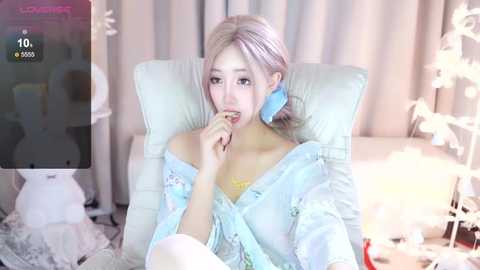 Media: Video of an Asian woman with platinum blonde hair, wearing a light blue robe, sitting on a white chair, biting her finger, in a softly lit room with white curtains and decorative ornaments.