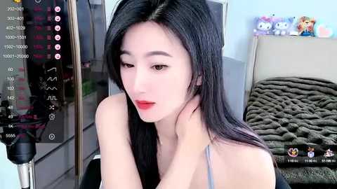 Media: Video of an East Asian woman with long black hair, fair skin, and red lipstick, wearing a light grey top, sitting in a room with a green blanket, toys, and a mirror.