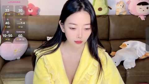 Media: Video of an East Asian woman with long black hair, wearing a yellow robe, seated on a brown leather couch. Background features plush toys, including a duck and a bear.