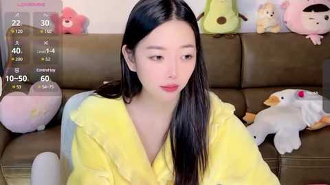 Media: A video of a young East Asian woman with long black hair, wearing a yellow robe, sitting on a brown leather couch. Surrounding her are plush toys, including a white duck and a green frog.