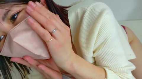 Media: Video of a young woman with light skin, wearing a beige mask and white knitted sweater, covering her mouth with her right hand.