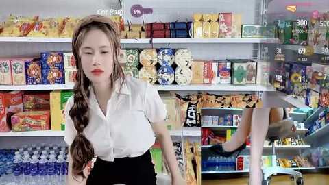 Media: Video of a Caucasian woman with long brown hair in braids, wearing a white blouse and black skirt, standing in a brightly lit supermarket aisle filled with colorful packaged snacks and beverages.