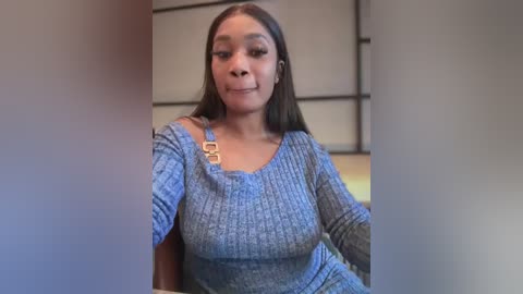 Media: Video of a young Black woman with long straight hair, wearing a blue ribbed sweater and a leather shoulder bag. She's indoors, with a blurred background featuring a white and beige patterned wall.