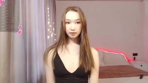 Media: Video of a young East Asian woman with long, straight brown hair, fair skin, and a slim build. She wears a black V-neck top. Background: white walls, bed, curtains, and red LED lights.