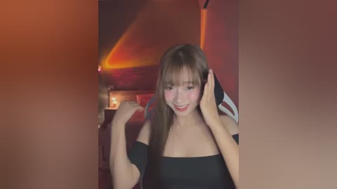 Media: Video of a young Asian woman with straight brown hair, fair skin, and a slender physique. She wears a black off-shoulder top, smiling with flushed cheeks, and playfully touches her face. Background features a dimly lit, warmly colored room with red and orange hues.