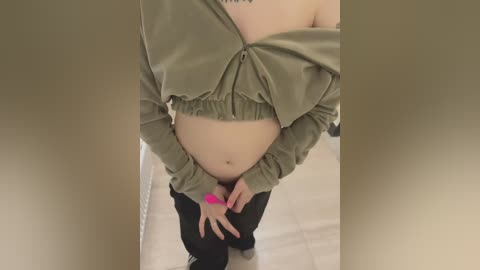 Media: Video of a pregnant woman in a cropped olive-green top, showing her bare midriff and belly. She wears black pants and a pink wristband, standing indoors on beige flooring.