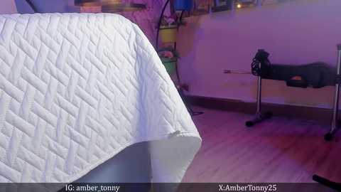 Media: Video of a neatly made bed covered in a white quilted bedspread in a softly lit room with a massage table and a partially visible person in the background.