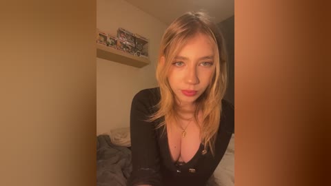 Media: A video of a fair-skinned, blonde woman with medium-length hair, wearing a black top, standing in a dimly lit bedroom with beige walls and a shelf with toys.