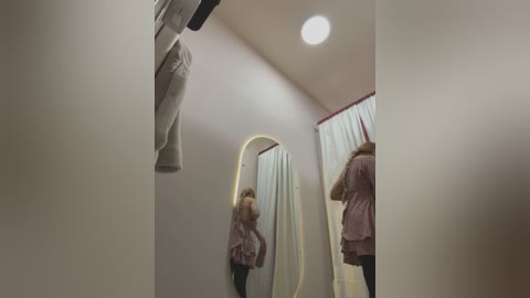 Media: Video of a beige-walled fitting room with a woman in a pink shirt and white pants, wearing a face mask, standing in front of a full-length mirror.